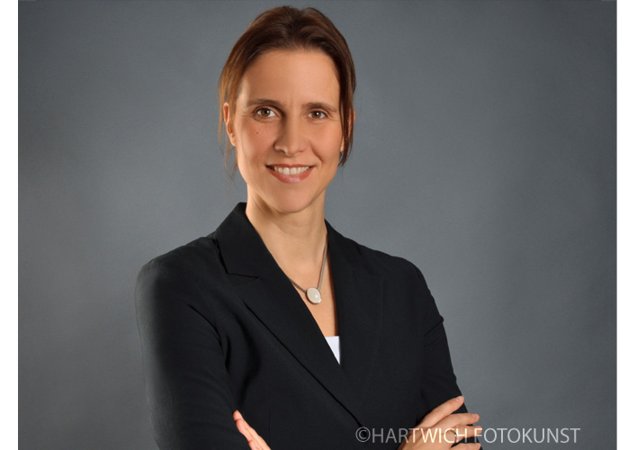 Professionelles Bewerbungsfoto einer Frau mit kurzen, dunkelbraunen Haaren. Sie trägt einen blauen Blazer über einer weißen Bluse mit gemustertem Kragen. Ihr Lächeln wirkt sympathisch und professionell. Der Hintergrund ist hellgrau.