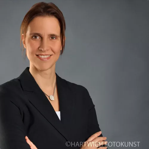 Professionelles Bewerbungsfoto einer Frau mit schulterlangem braunem Haar. Sie trägt einen dunklen Blazer über einem weißen Oberteil sowie eine dezente Halskette mit rundem Anhänger. Mit freundlichem Lächeln und aufrechter Haltung blickt sie selbstbewusst in die Kamera. Der Hintergrund ist neutral grau, passend für eine seriöse Darstellung.