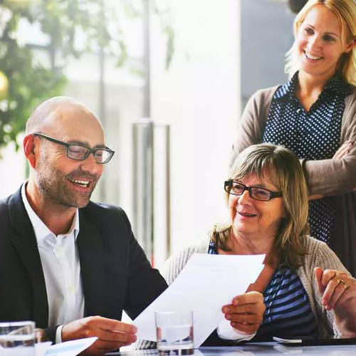 Drei Personen in einem modernen, lichtdurchfluteten Büro in einer freundlichen Besprechung. Ein glatzköpfiger Mann mit Brille und dunklem Anzug hält ein Dokument und lacht. Neben ihm sitzt eine ältere Frau mit Brille und schulterlangem Haar, ebenfalls lächelnd. Hinter ihnen steht eine blonde Frau in einer Strickjacke und schaut lächelnd zu. Alle wirken entspannt und engagiert. Auf dem Tisch stehen Gläser mit Wasser.