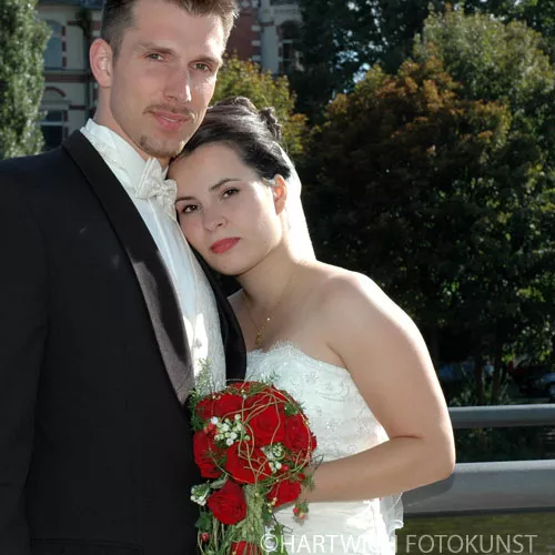 Hochzeitsfoto eines frisch verheirateten Paares im Freien. Der Bräutigam, ein junger Mann mit kurzen Haaren und leichtem Bart, trägt einen eleganten schwarzen Anzug mit weißem Hemd und Fliege. Die Braut, eine junge Frau mit dunklem, hochgestecktem Haar, lehnt sich an ihn und hält einen Strauß roter Rosen. Sie trägt ein weißes Hochzeitskleid mit schulterfreiem Schnitt. Im Hintergrund sind grüne Bäume und ein historisches Gebäude zu sehen. Die Abendsonne wirft ein warmes Licht auf die Szene.