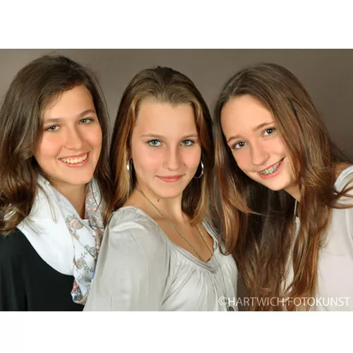Porträtfoto von drei jungen Frauen mit langen Haaren, die fröhlich in die Kamera lächeln. Sie stehen eng beieinander vor einem neutralen, hellbraunen Hintergrund. Die linke trägt ein schwarzes Oberteil mit einem weißen Schal, die mittlere ein hellgraues Oberteil mit V-Ausschnitt und einer Silberkette, die rechte ein weißes Oberteil und hat eine Zahnspange. Die Aufnahme strahlt Wärme und Verbundenheit aus.